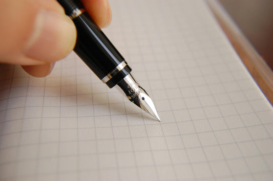 Close-up of a hand holding a sleek black fountain pen with a silver nib, writing on a grid paper notebook. Perfect for highlighting precision writing tools, high-quality pens, and professional writing instruments."